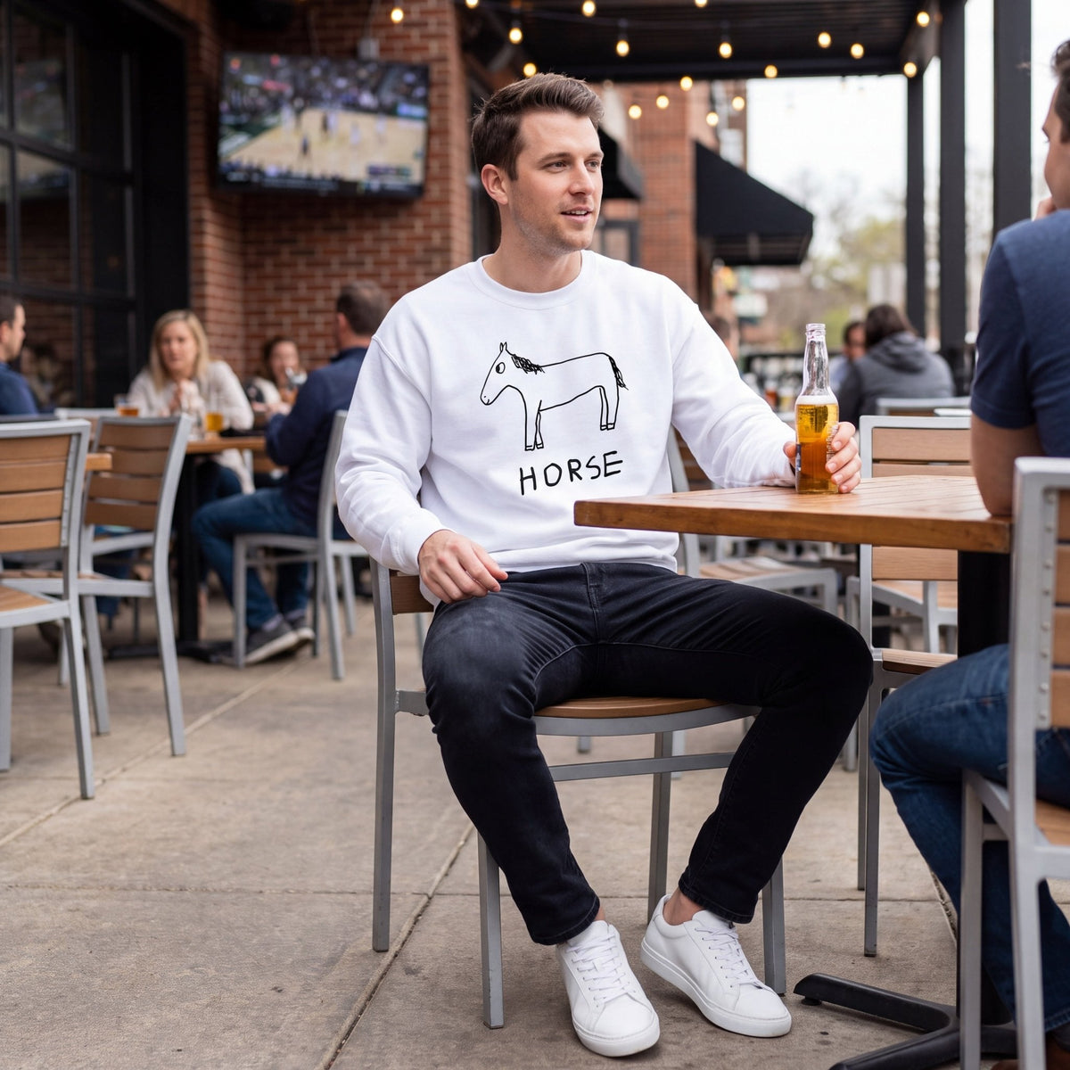 A man sitting at an outdoor patio table wearing a white crewneck sweatshirt with a black line art horse graphic and the word 'HORSE' printed below it, paired with black jeans and white sneakers.