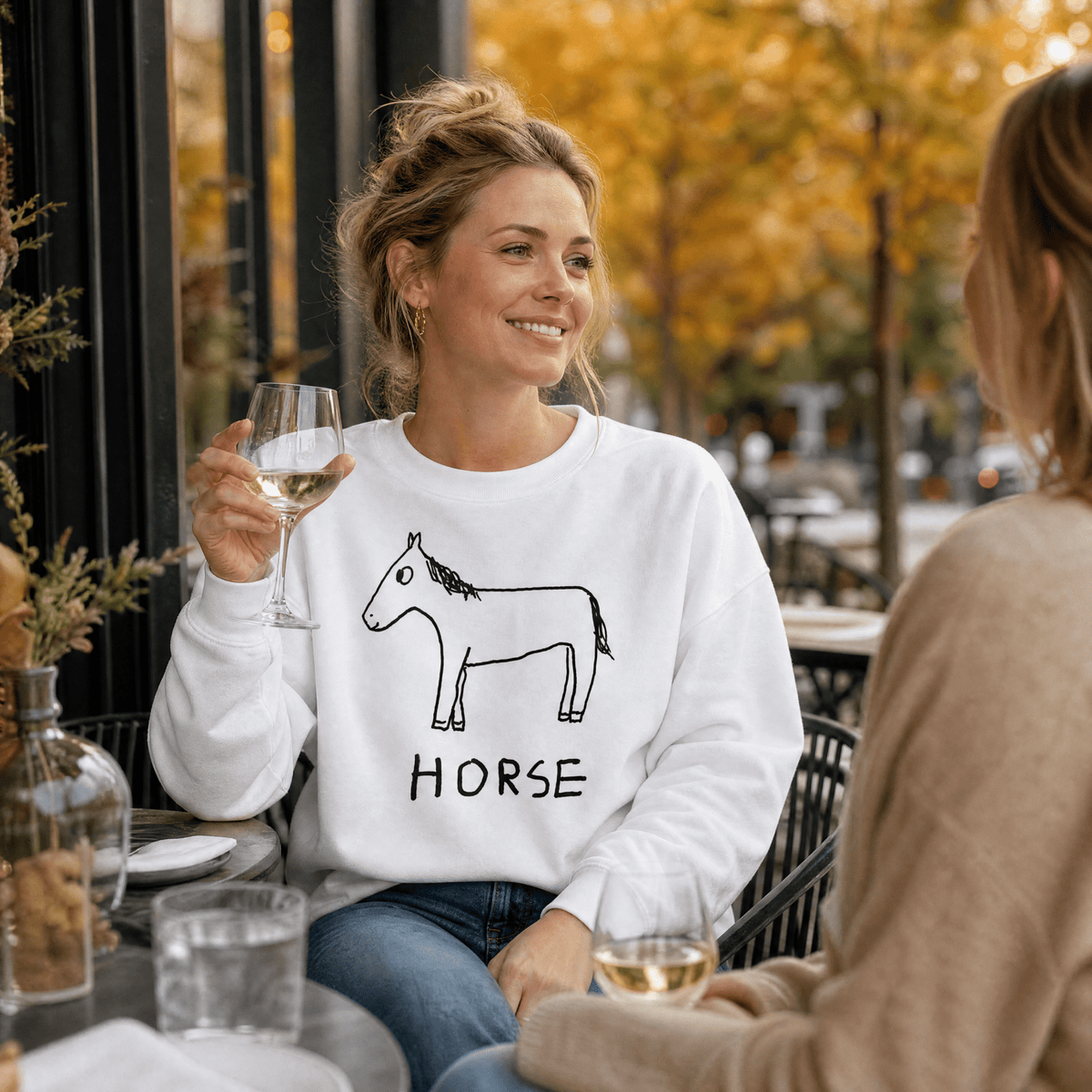 A woman sitting at an outdoor cafe table wearing a white crewneck sweatshirt with a black line art horse graphic and the word 'HORSE' printed below it. She is holding a glass of white wine and smiling at her conversation partner.