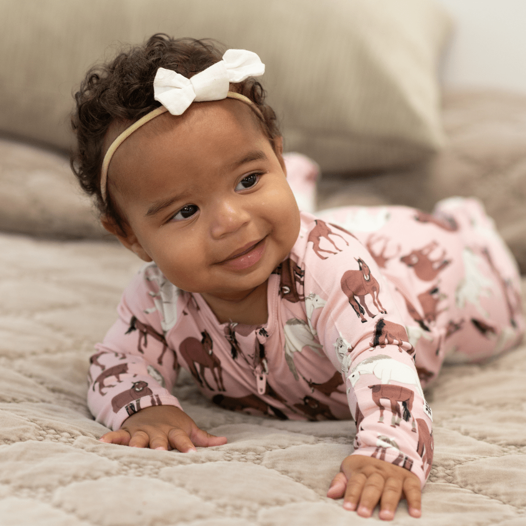 A baby lying on a quilted blanket wearing a pink long-sleeve one-piece pajama with a horse print and a white bow headband.