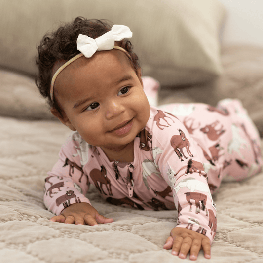 A baby lying on a quilted blanket wearing a pink long-sleeve one-piece pajama with a horse print and a white bow headband.