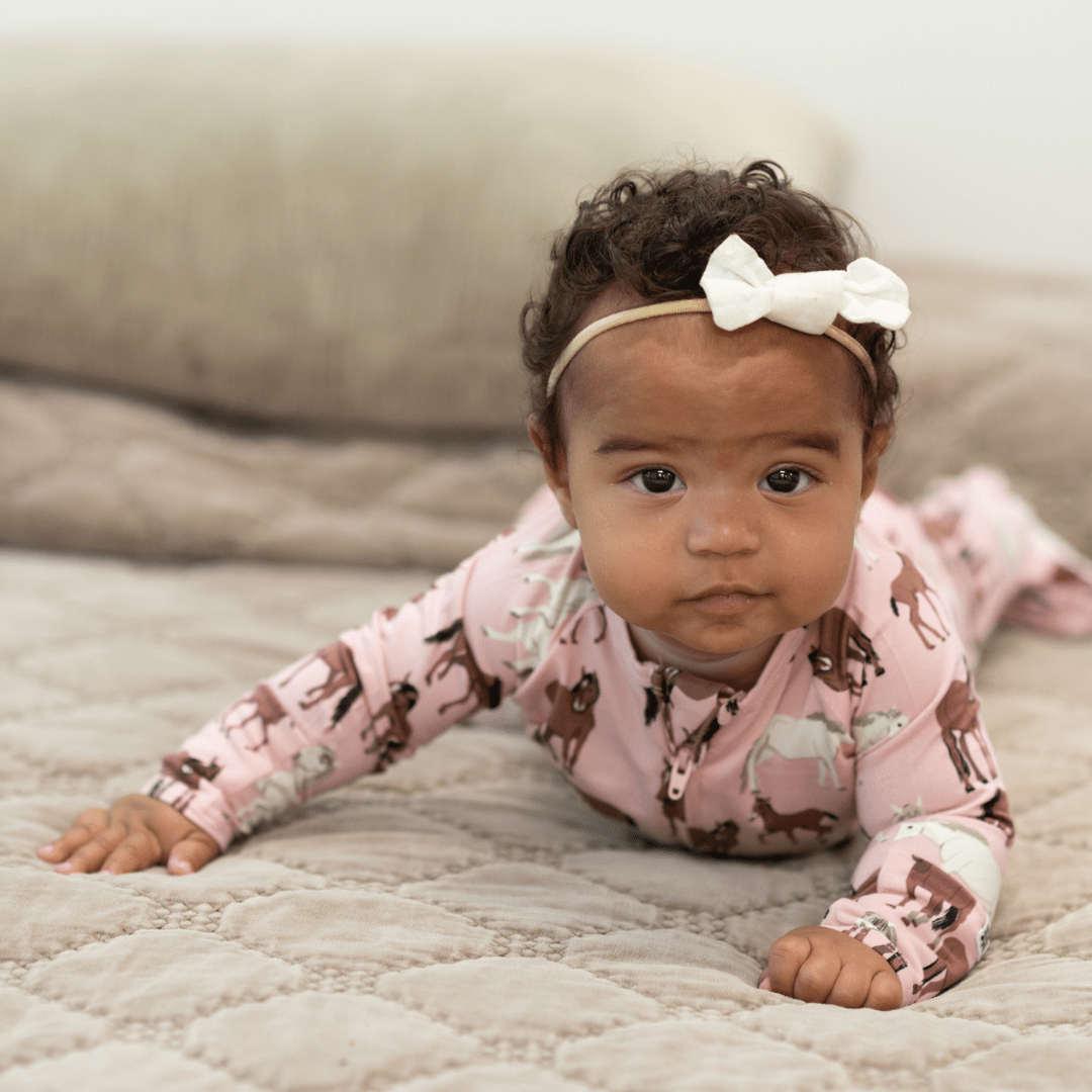 Baby wearing a pink outfit with animal prints and a white headband, lying on a beige surface.